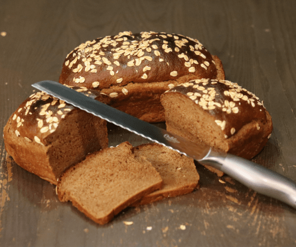 two loaves of homemade brown pumpernickel bread with rolled oats on top, one loaf is whole and the other loaf is sliced in half with 2 slices of bread showing with a metal bread knife leaning on one half of the sliced bread